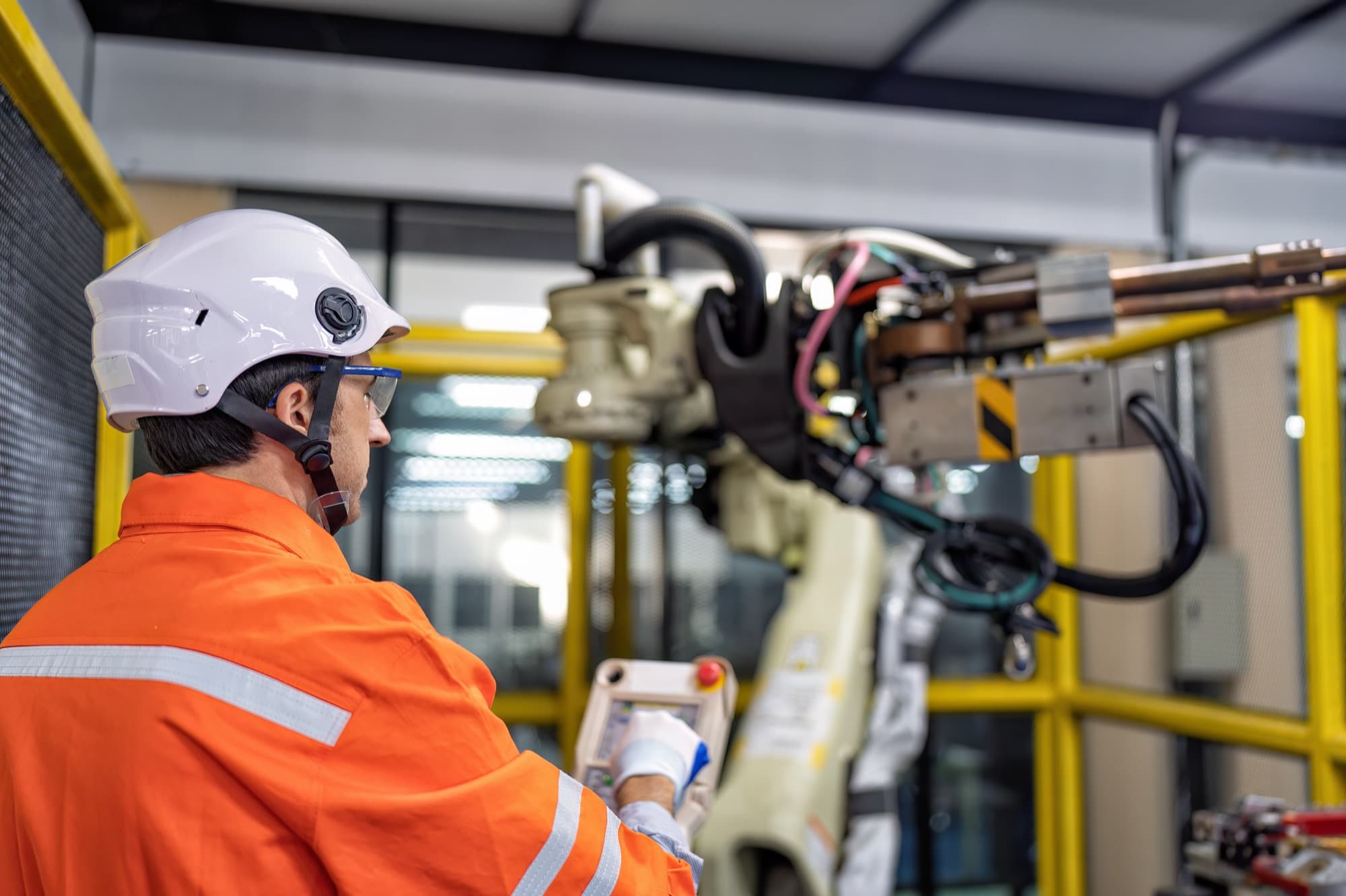 Technicien casqué pilotant un bras robot depuis le boîtier de commande, derrière la grille de la cellule