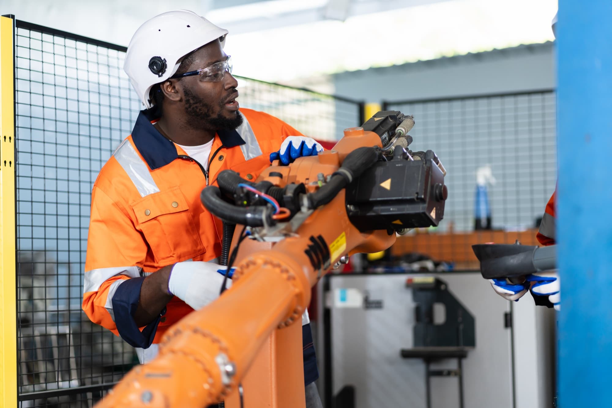 Technicien en gilet haute visibilité inspectant le poignet d'un bras robot orange dans sa cellule grillagée