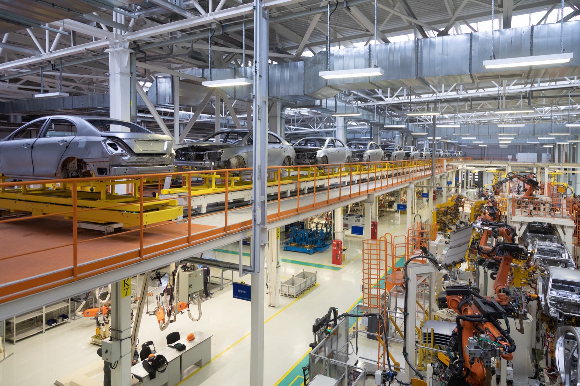Vue plongeante sur un atelier automobile, caisses sur convoyeur aérien et rangée de robots orange