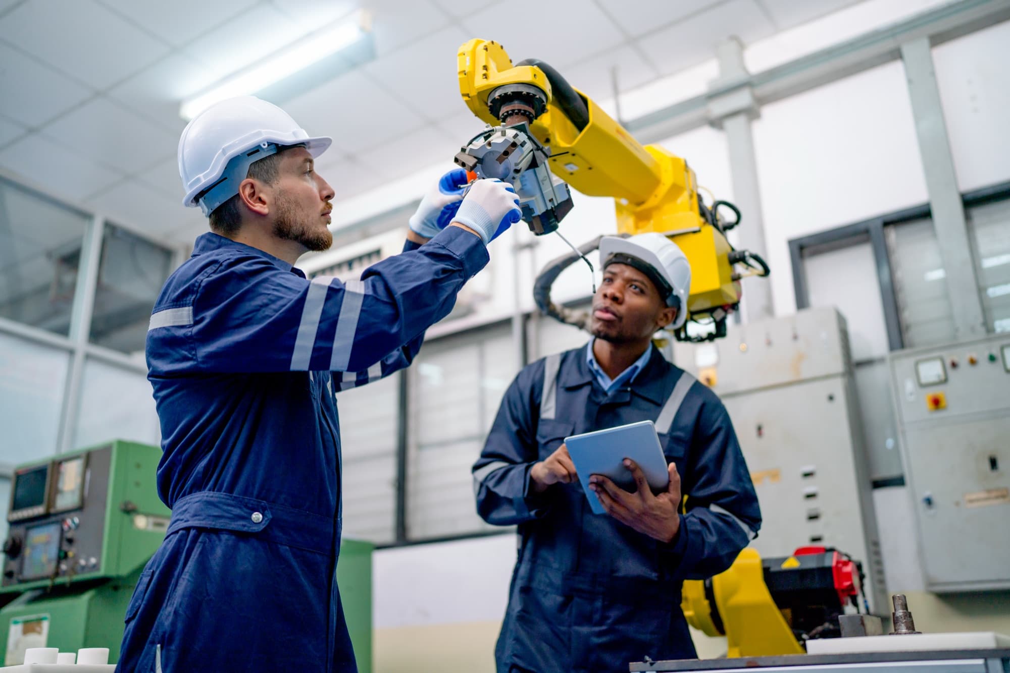 Deux techniciens en combinaison bleue examinant la pince d'un robot jaune dans un atelier de production