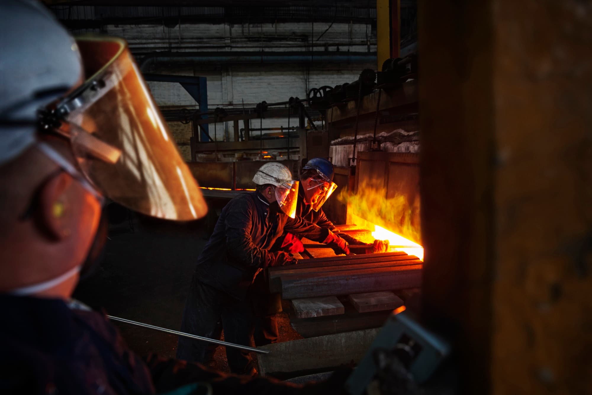 Ouvriers protégés par des écrans faciaux poussant des barres d'acier devant un four incandescent