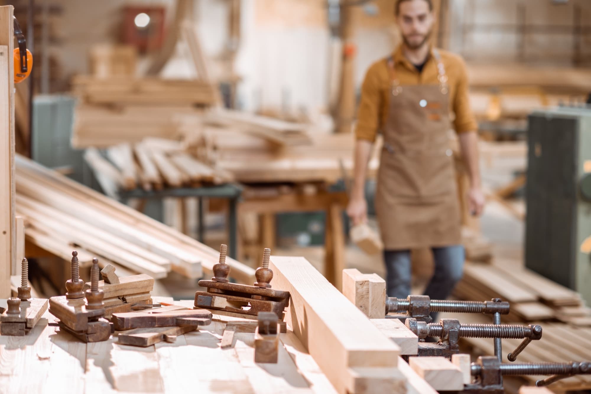 Serre-joints et pièces de bois en premier plan sur un établi, menuisier en arrière-plan dans l'atelier