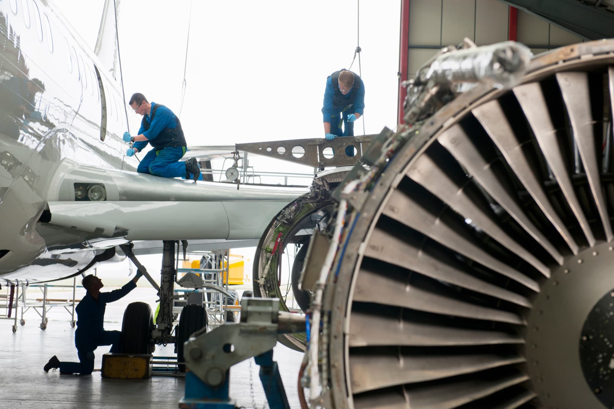 Trois mécaniciens intervenant sur la voilure et le train d'un avion en hangar, réacteur déposé au premier plan