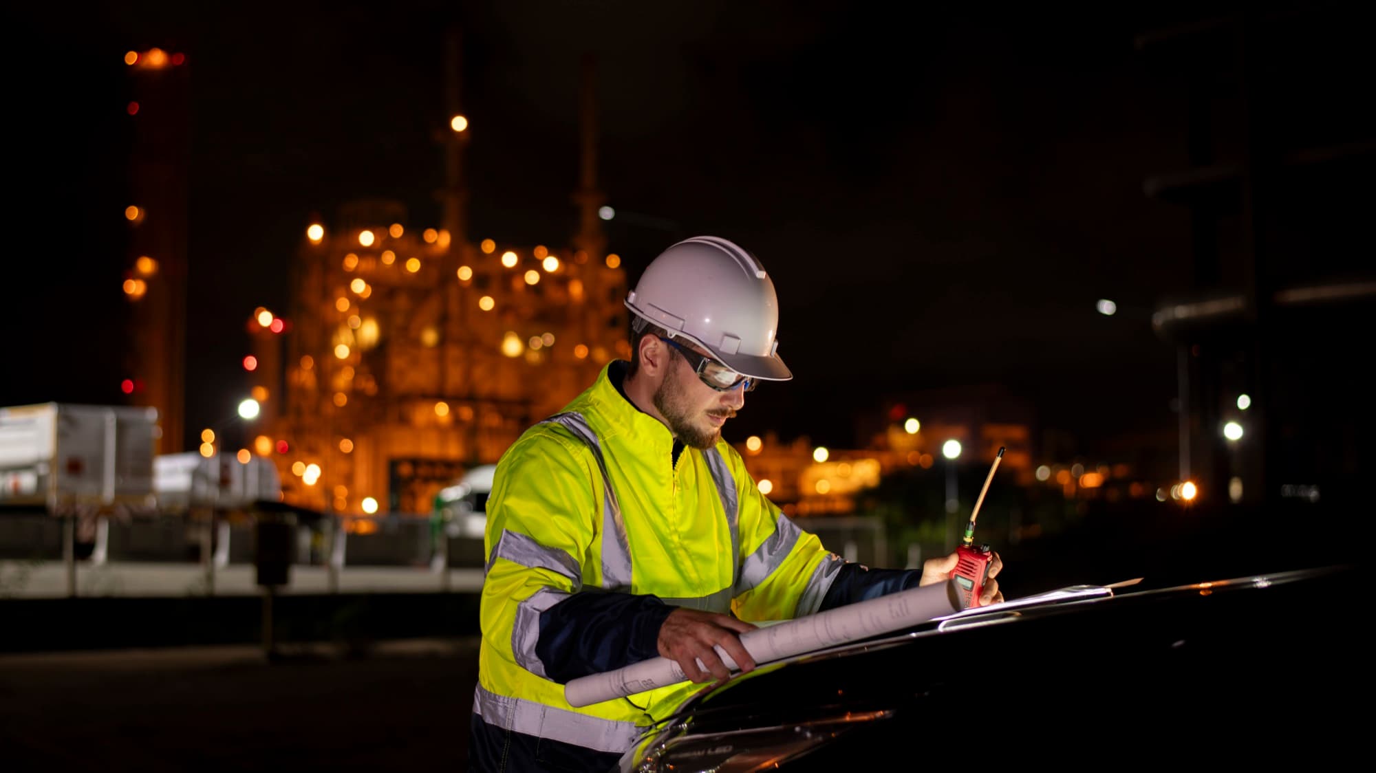 Technicien en gilet haute visibilité annotant des plans de nuit devant une usine pétrochimique illuminée