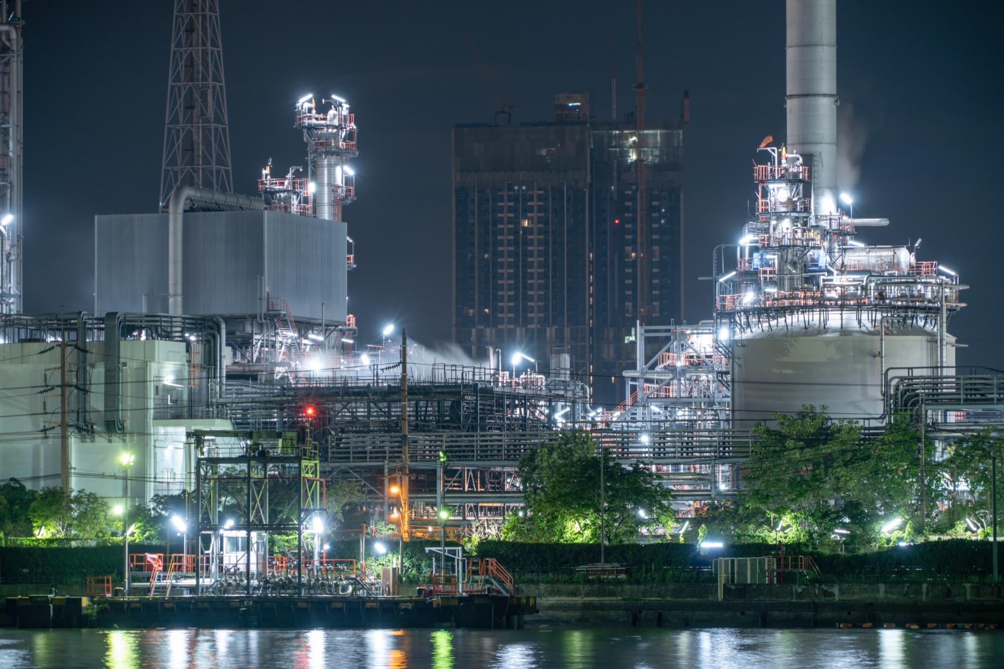 Usine pétrochimique éclairée de nuit au bord d'un fleuve, réservoirs et racks de tuyauterie