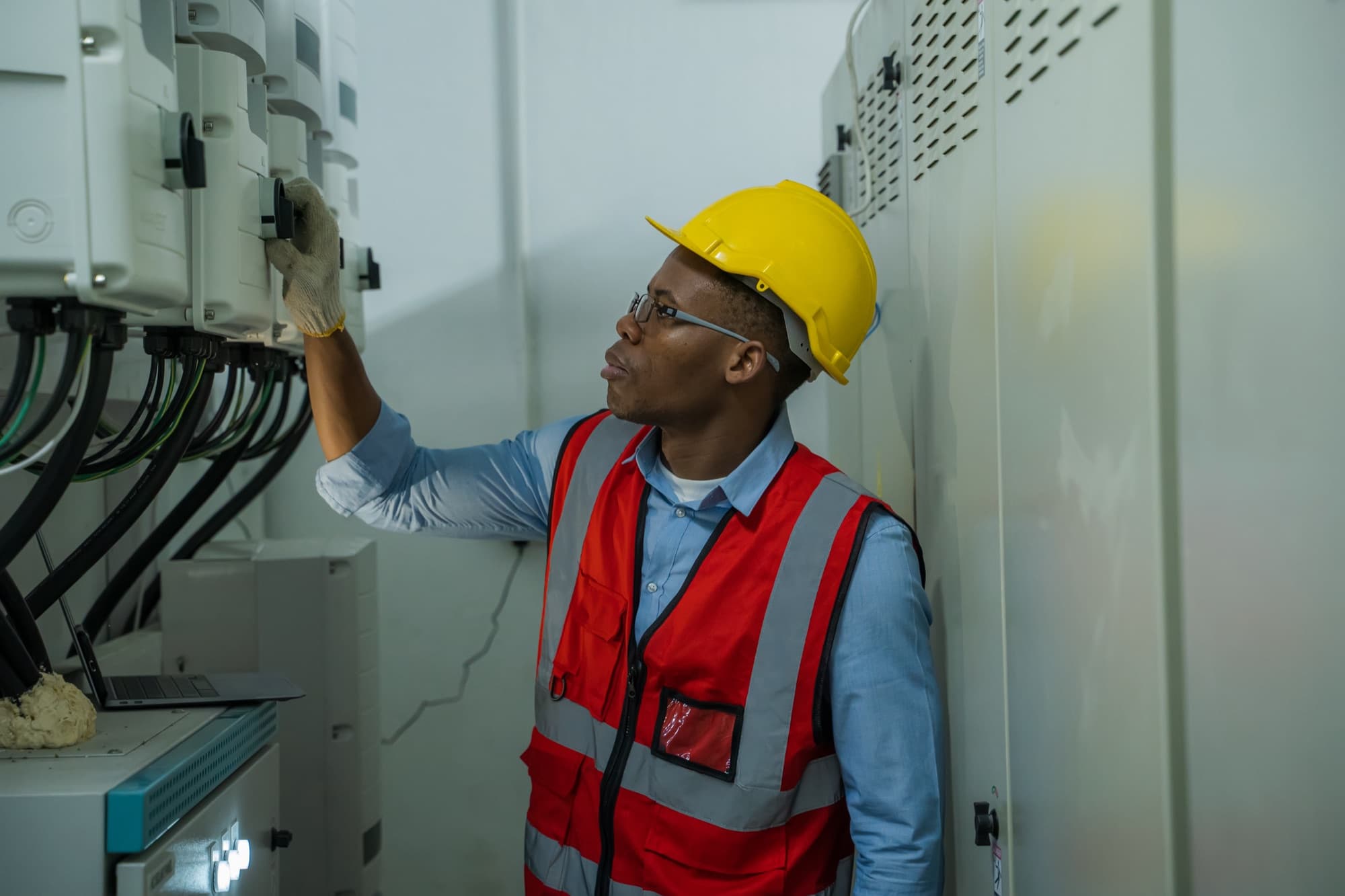 Technicien en gilet rouge et casque jaune vérifiant une rangée de coffrets électriques dans un local de distribution