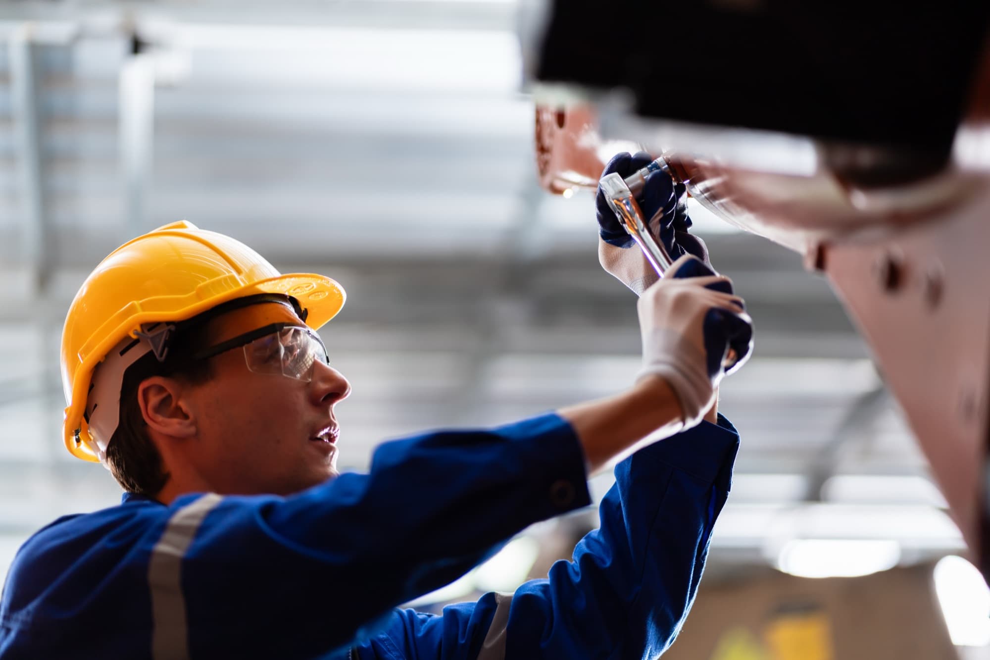 Technicien en casque jaune serrant un assemblage à la clé à cliquet au-dessus de sa tête dans une halle de production