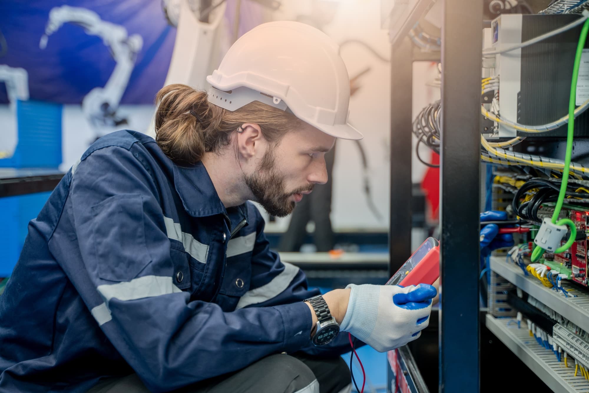 Technicien accroupi mesurant au multimètre dans une armoire d'automatisme ouverte, bras robotisé en arrière-plan