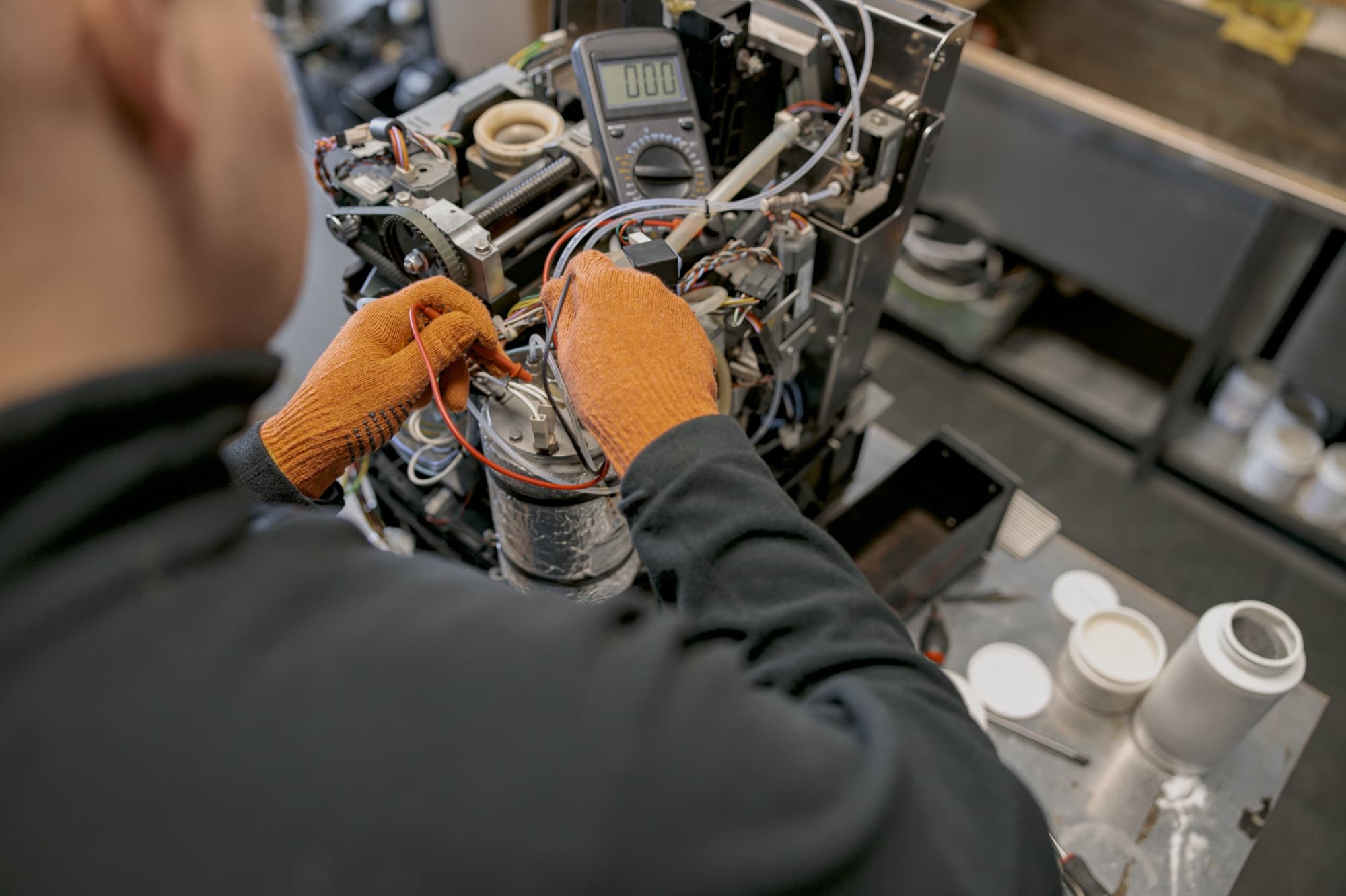 Réparateur en gants orange intervenant sur les entrailles d'une machine à café, multimètre branché sur l'établi