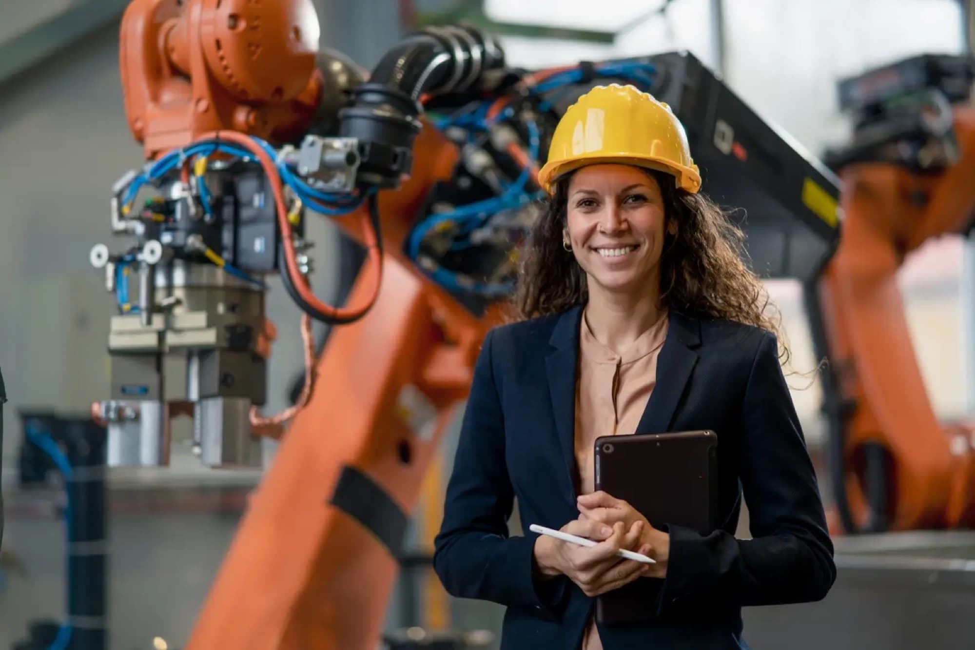 Ingénieure en veste de tailleur et casque de chantier, tablette à la main, devant des robots industriels orange