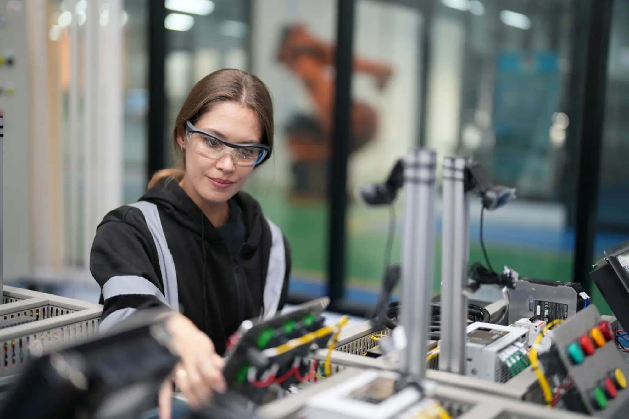 Technicienne câblant un banc d'automatisme, borniers, automate et boutons poussoirs, un robot en arrière-plan