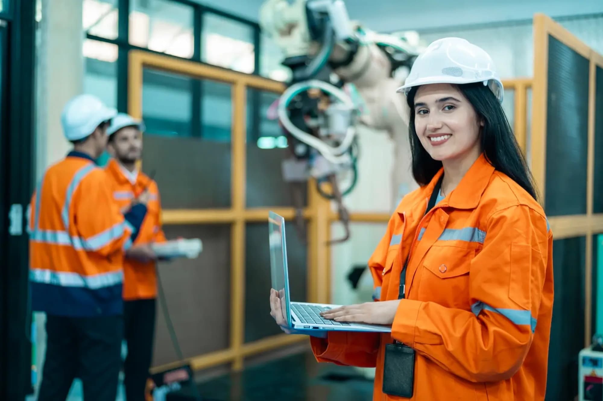 Technicienne ordinateur portable ouvert devant une cellule robotisée, deux collègues en discussion derrière elle