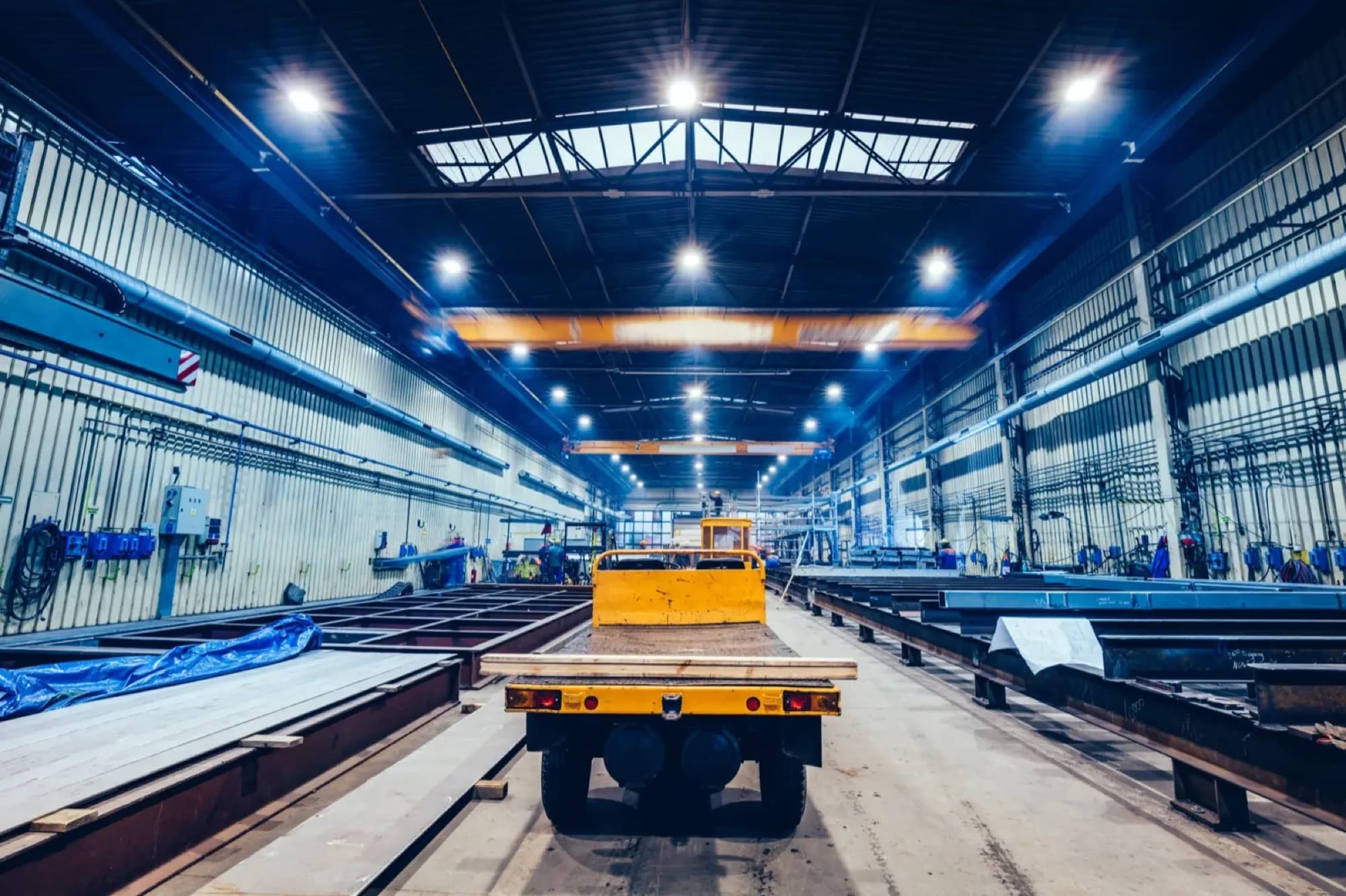 Halle de construction métallique, poutres et tôles alignées au sol, plateau automoteur jaune sous les ponts roulants