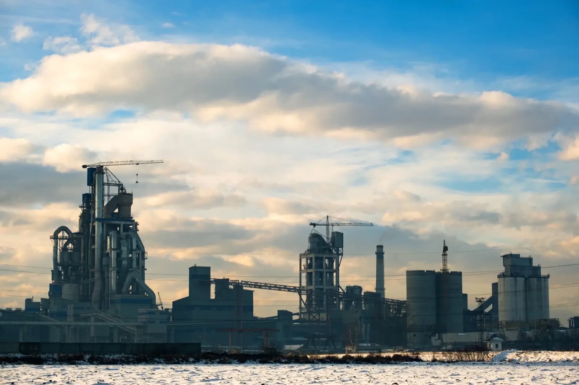Cimenterie en hiver, tour de préchauffage, silos et grues à tour derrière un champ enneigé