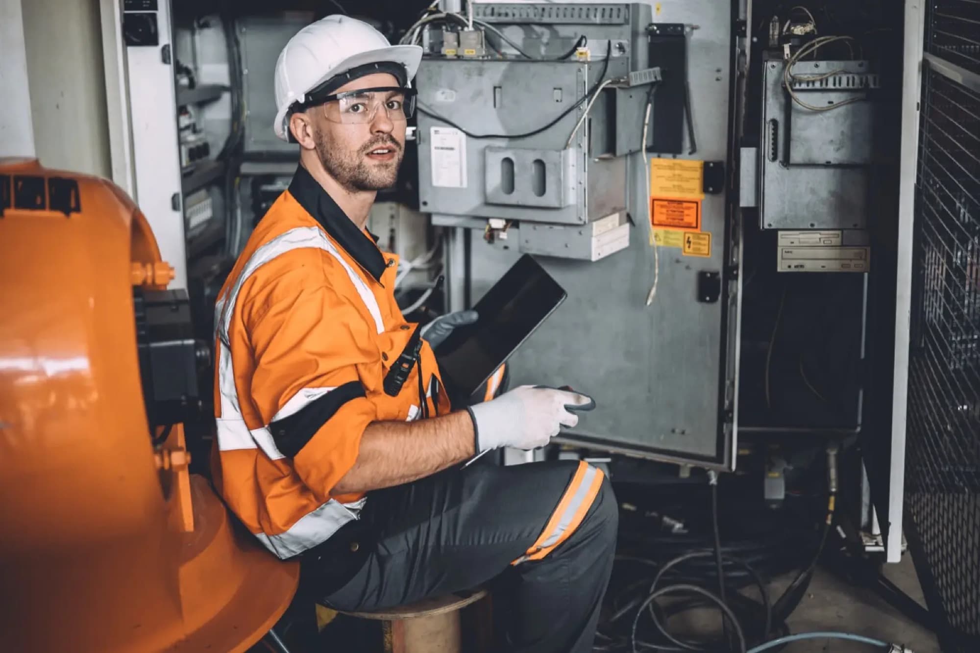 Technicien assis tablette en main devant une armoire électrique ouverte, à côté d'un robot industriel orange