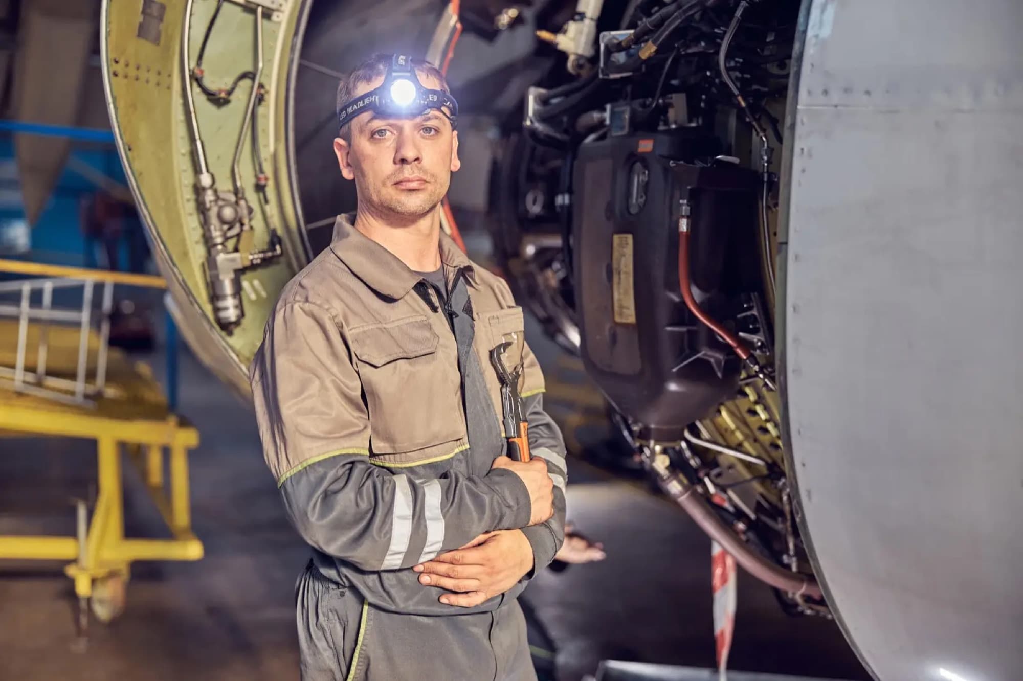 Mécanicien à lampe frontale, clé à molette sous le bras, devant un réacteur d'avion capots ouverts dans un hangar