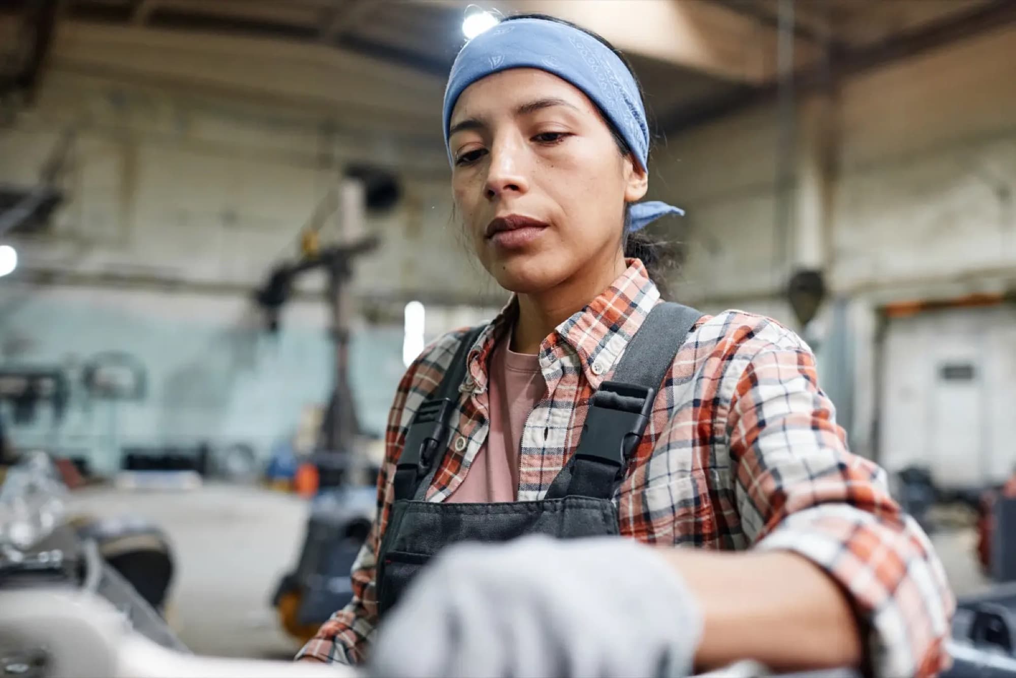 Opératrice en bandana et salopette, gants de travail, penchée sur une machine dans un atelier industriel