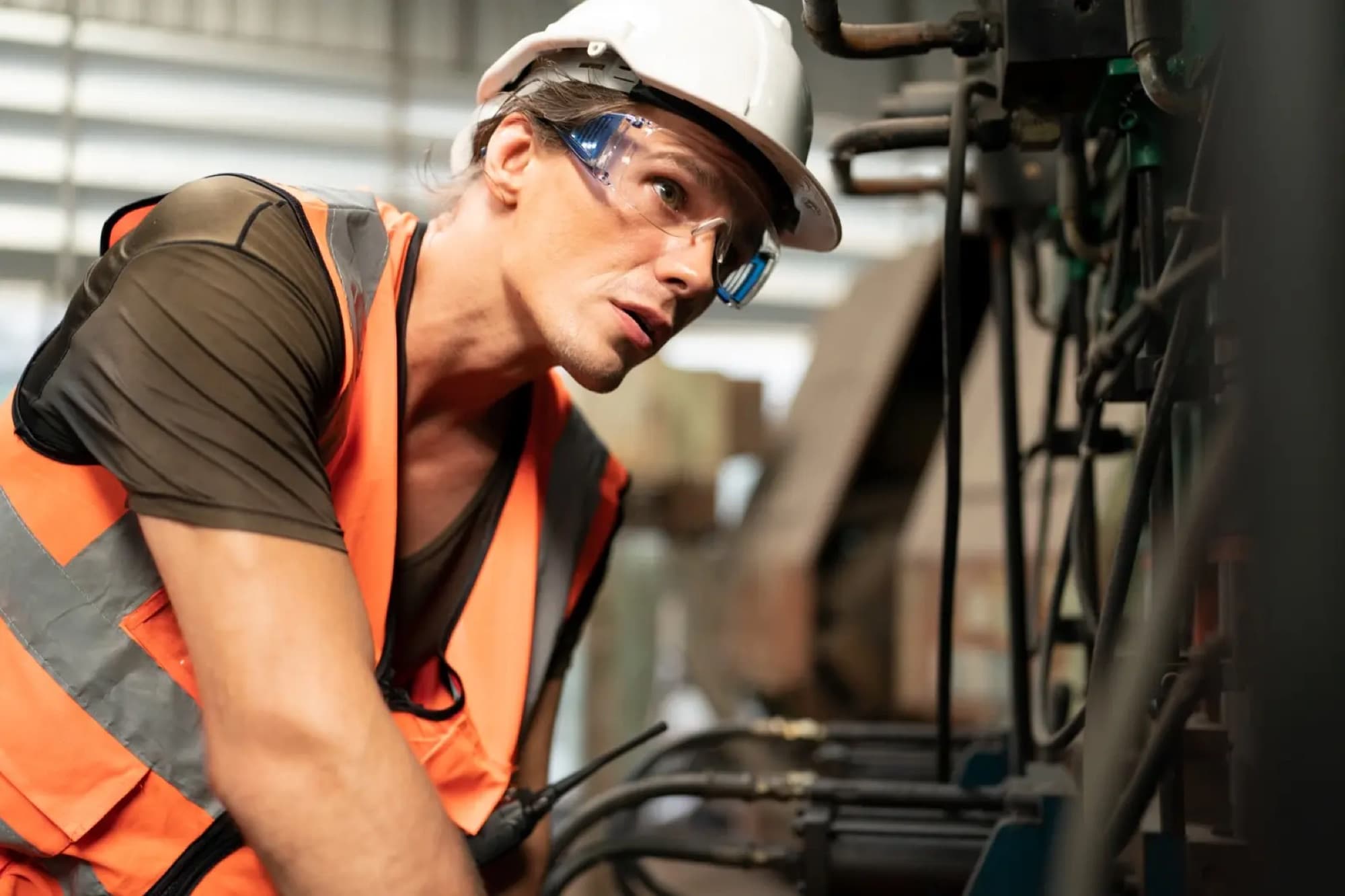 Technicien en casque blanc et gilet orange inspectant de près une machine garnie de flexibles et de vérins