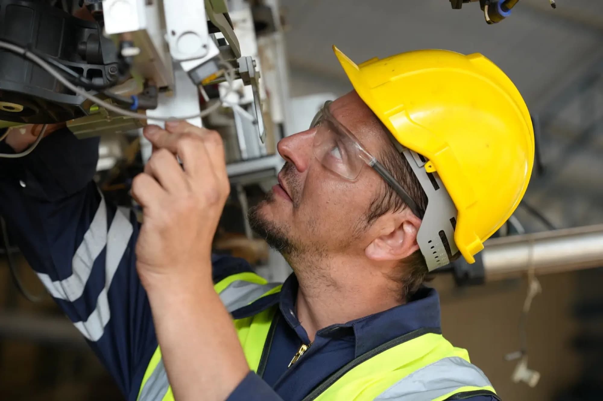 Technicien en casque jaune intervenant bras levés sur des capteurs et des vérins en partie haute d'une machine