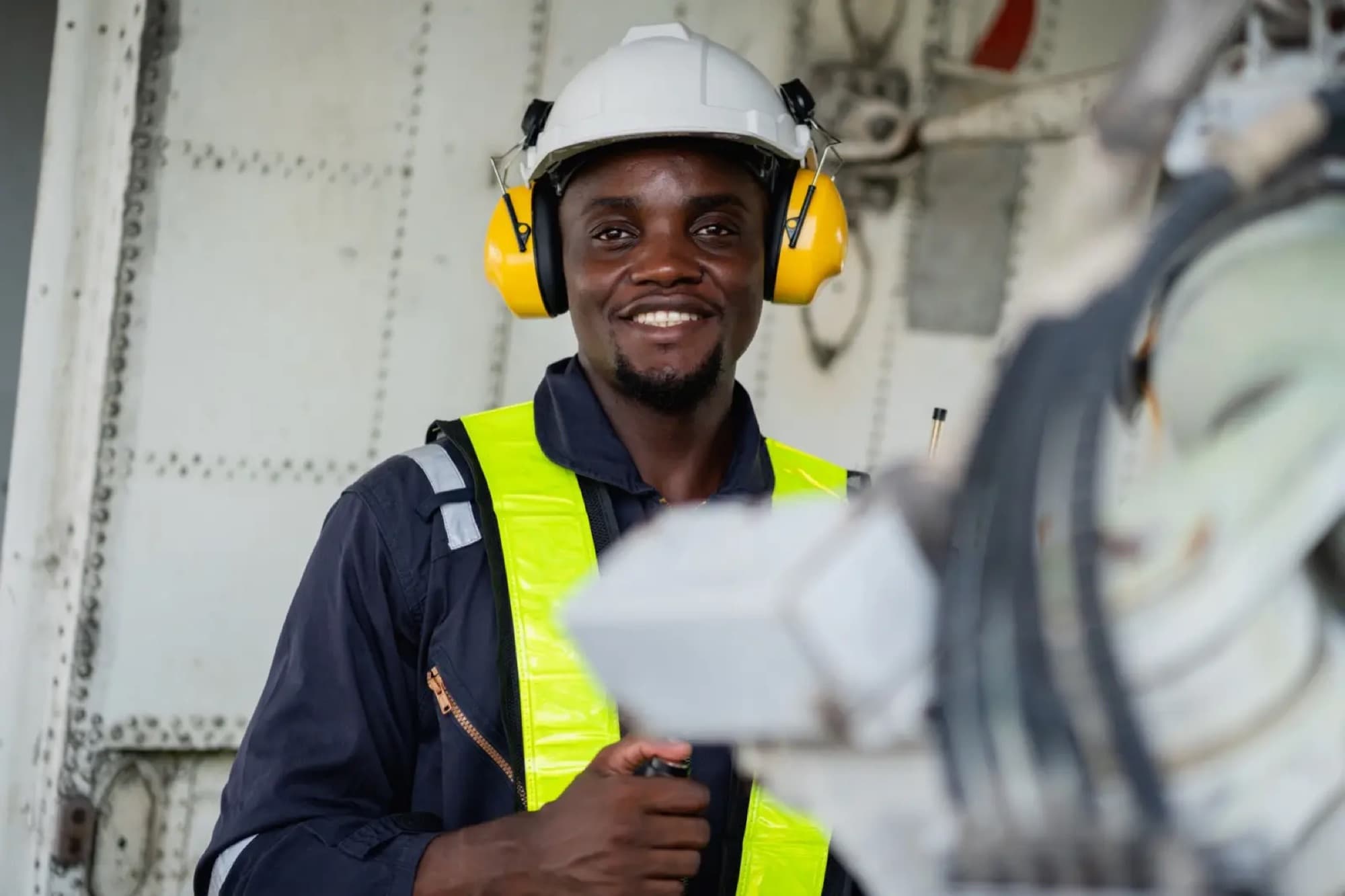 Technicien en casque et protections auditives souriant près d'une machine, devant une paroi métallique rivetée