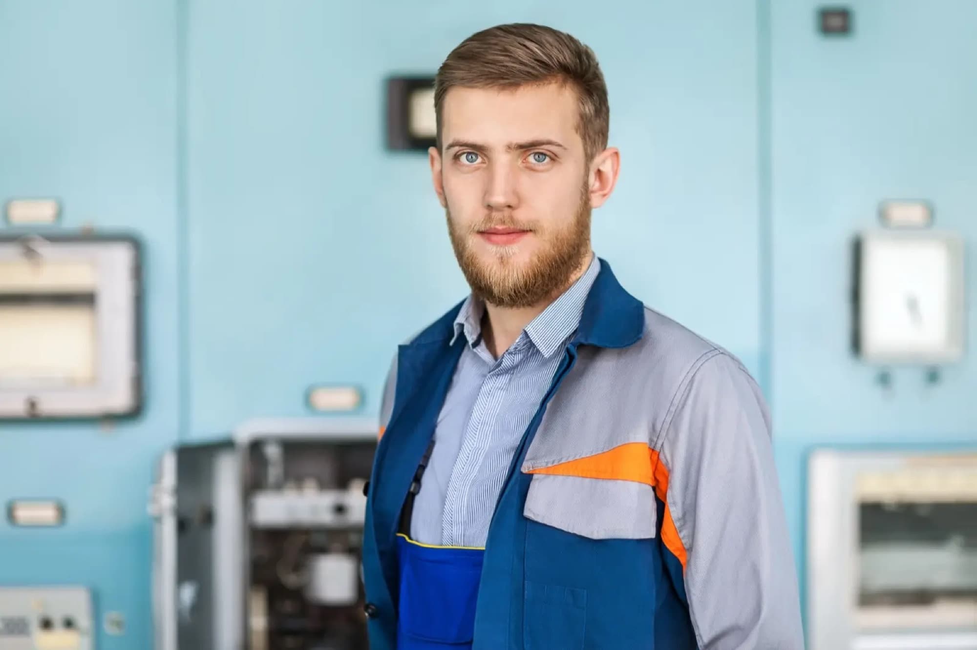 Portrait d'un technicien en veste de travail devant les coffrets électriques muraux d'un local technique