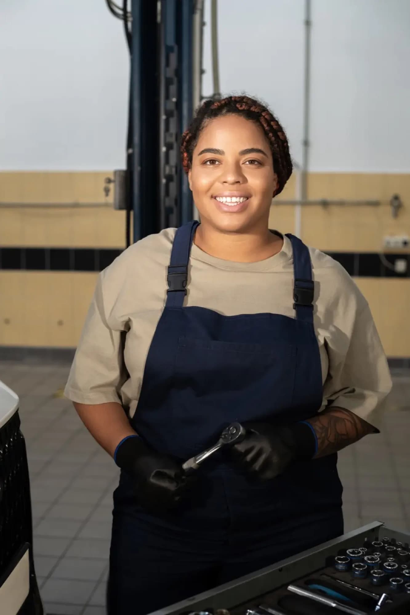 Mécanicienne souriante, cliquet en main, devant sa servante à outils dans un garage automobile à pont élévateur