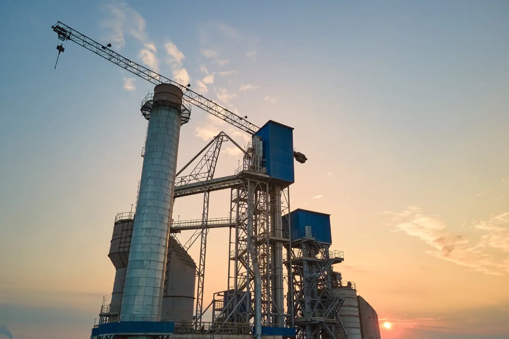Silos et convoyeurs inclinés d'une installation cimentière au coucher du soleil, surmontés d'une grue à tour
